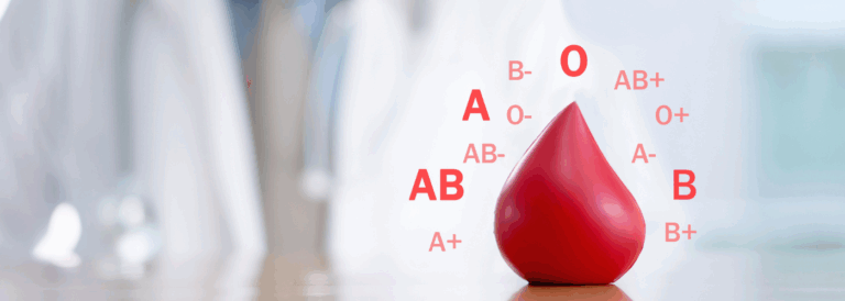 Imagen de una gota de sangre roja con diferentes grupos sanguíneos (A, B, AB y O, positivos y negativos) representados alrededor, sobre un fondo desenfocado en tonos claros.