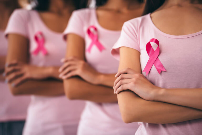 Grupo de mujeres con remeras rosas y lazos del mismo color en el pecho, símbolo de la lucha contra el cáncer de mama, de pie con los brazos cruzados en señal de unión y apoyo.