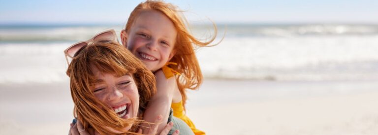 Madre e hija pelirrojas sonríen y se abrazan en la playa, con el mar de fondo y la brisa moviendo su cabello.
