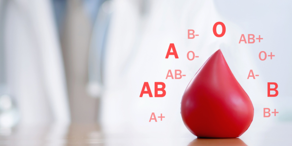 Imagen de una gota de sangre roja con diferentes grupos sanguíneos (A, B, AB y O, positivos y negativos) representados alrededor, sobre un fondo desenfocado en tonos claros.