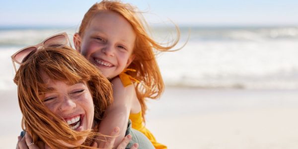 Madre e hija pelirrojas sonríen y se abrazan en la playa, con el mar de fondo y la brisa moviendo su cabello.